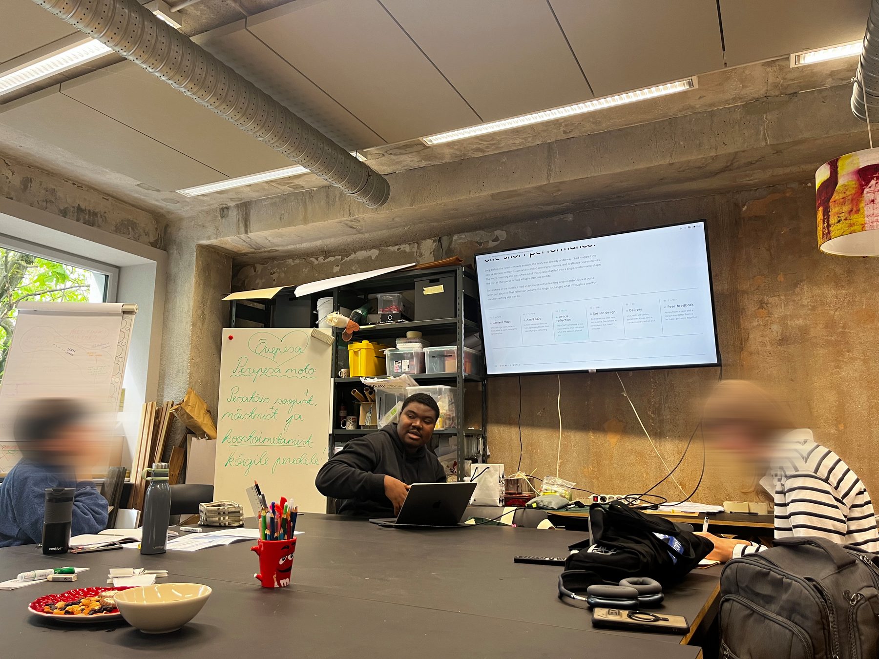 The micro-teaching session in progress: Faruk seated at a long table with a laptop, the portfolio page shown on a large wall-mounted screen, two participants partially visible in the foreground.